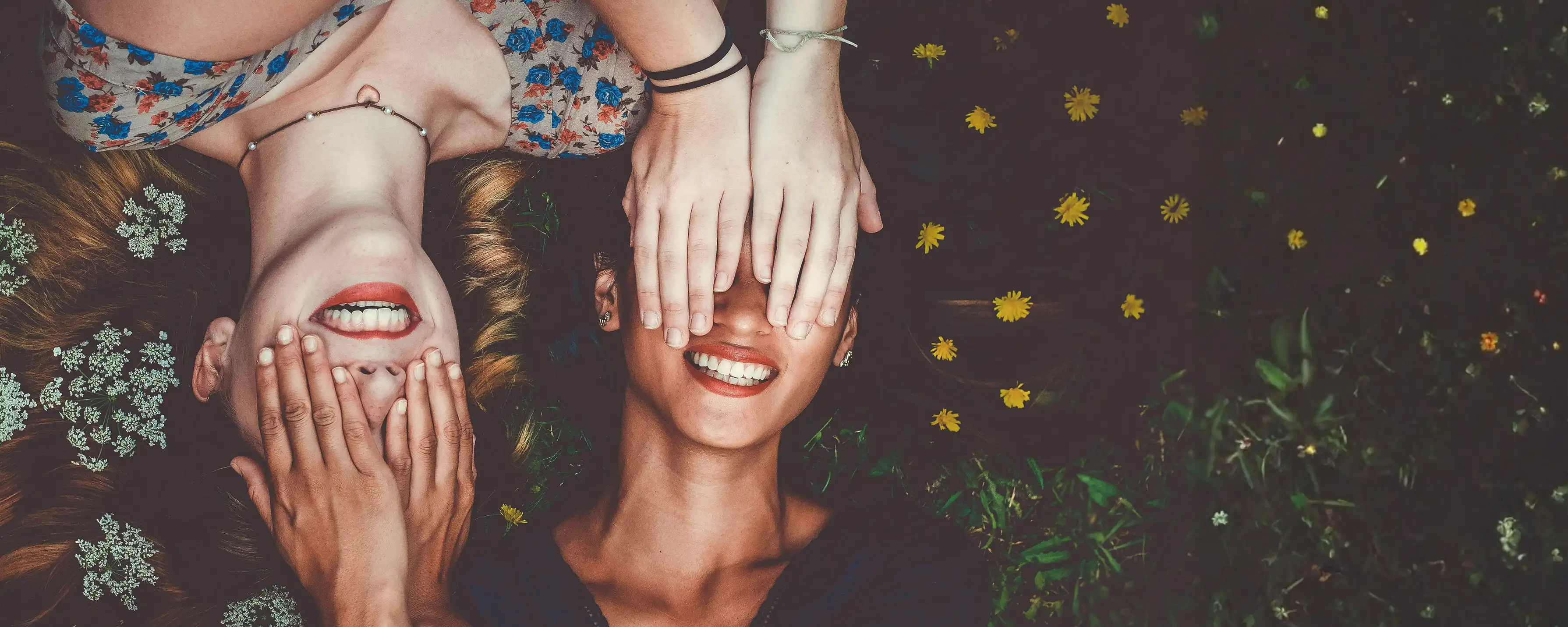 Zwei Freundinnen liegen gemeinsam auf der Wiese, halten sich gegenseitig die Augen zu und lachen.